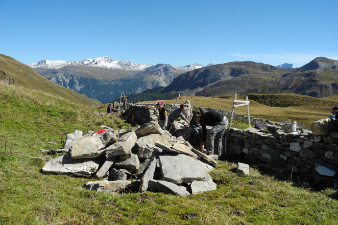 Drei Menschen beigen Steine von einem Haufen zu einer Steinmauer auf.