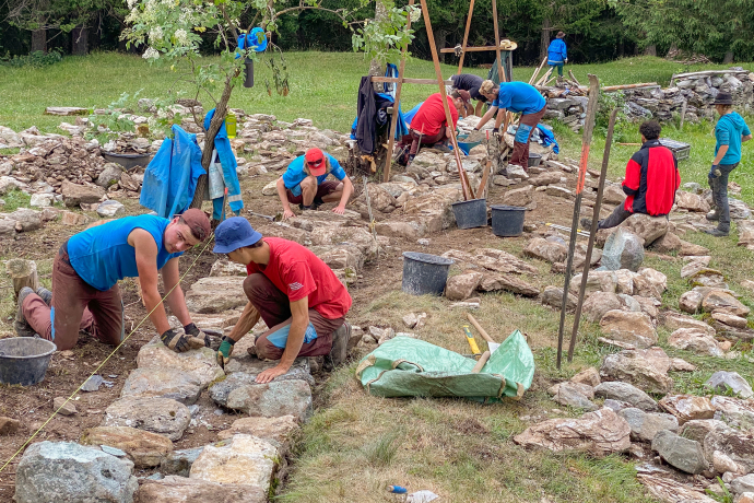 Trockenmauer-Kurs im Naturpark Beverin. Junge Männer und Frauen arbeiten teils kniend an einer Steinmauer auf einer Wiese.
